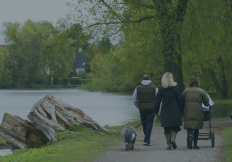Three people walking on a park with a river on the side; one is pushing a stroller and another one has a dog on a leash.